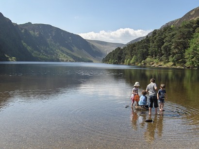 Waterside view of Glendalough's Upper Lake, hemmed in on three sides by high hills. In foreground is a group of four children, paddling in the shallows.