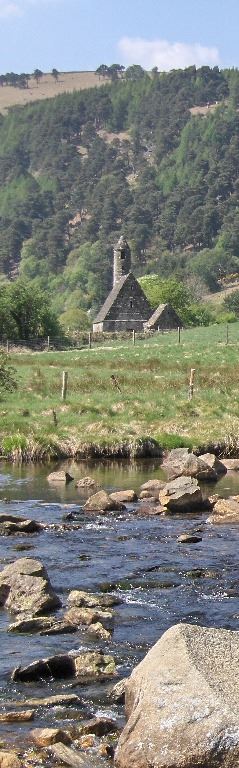 View of St Kevin's church at Glendalough in mid-distance, across rocky river and fields, with mountains and sky as backdrop.