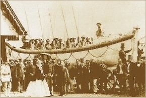 Photo taken at the launch of new Wicklow lifeboat in mid-180s. Mariners standing/sitting in the boat. Audience includes woman in crinoline and man in high top hat, other dignitaries, and several less-privileged local people.